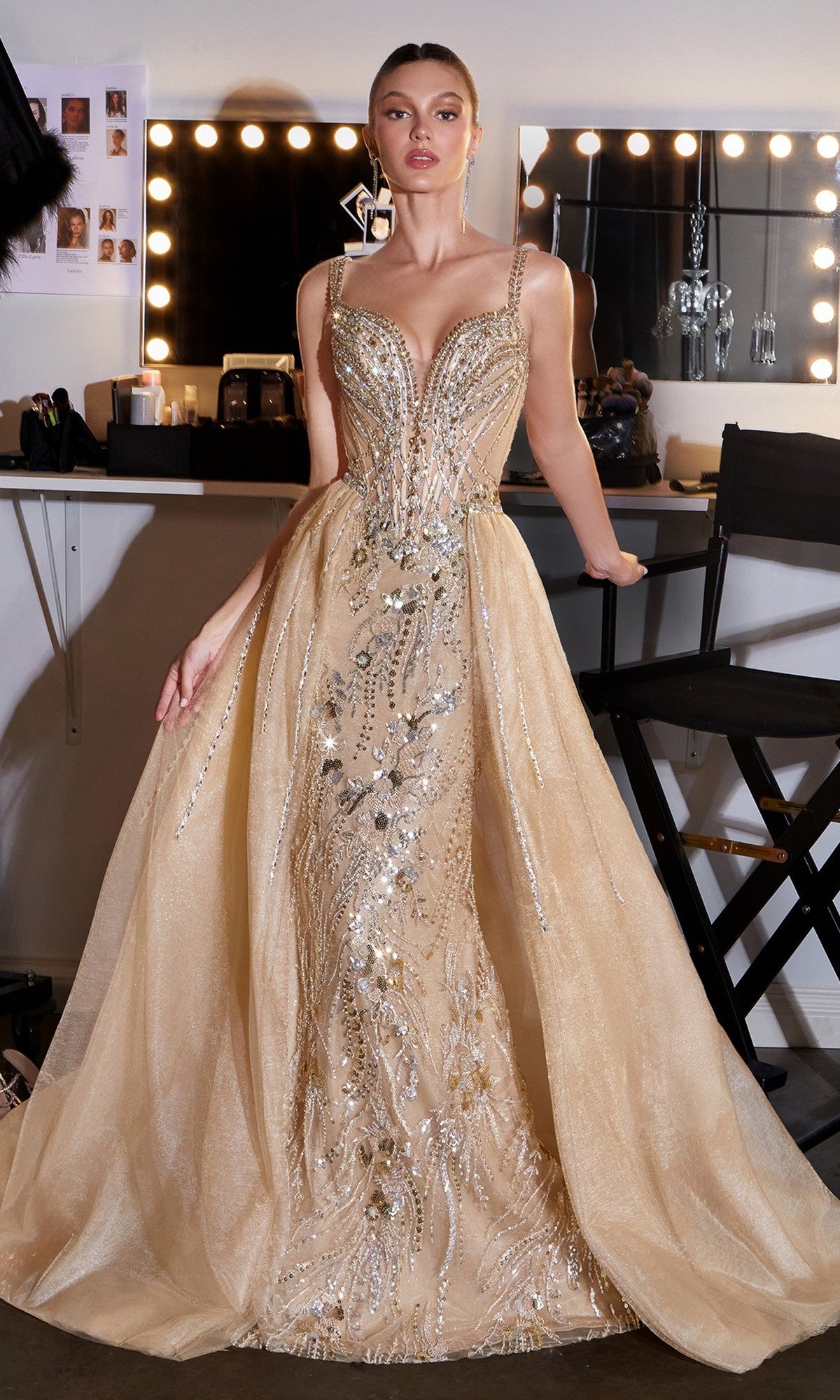 A woman stands in a dressing room wearing the Ladivine CDS570 Long Prom Dress, featuring gold crystal embellishments, a fitted bodice, sheer panels, and flared skirt. Makeup lights and cosmetics in the background complete this formal event look.